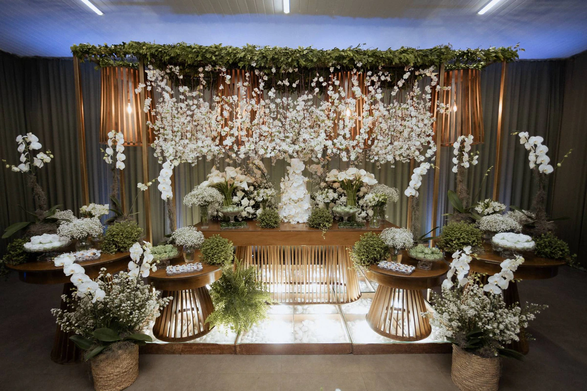 Mesa principal com estrutura dourada, cascata de flores brancas, luminárias ripadas e mesas ripadas sobre piso de vidro iluminado