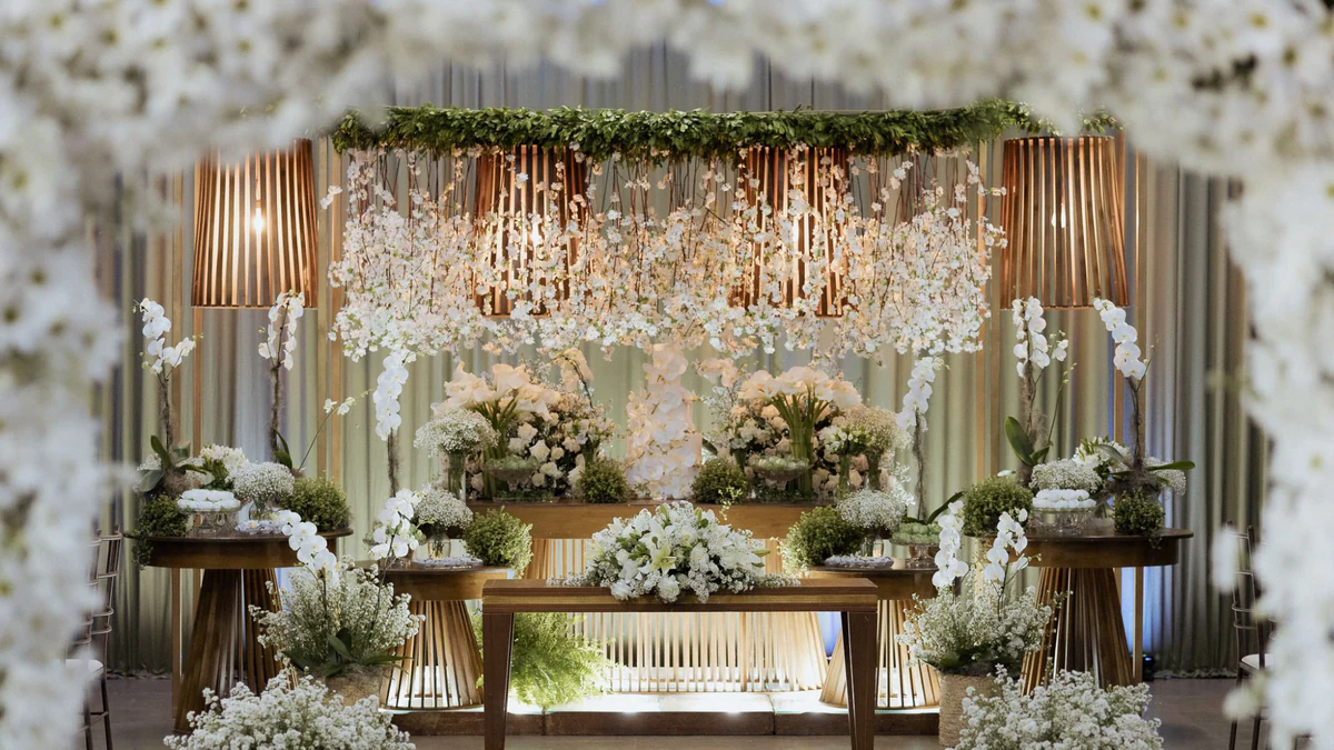 Mesa principal vista pelo arco de margaridas, com cascata de flores brancas, orquídeas, luminárias ripadas e altar à frente