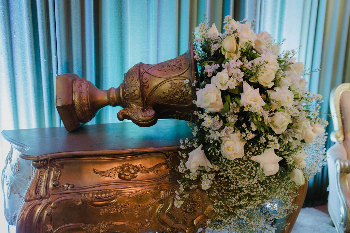 Vaso dourado tombado sobre aparador dourado, com cascata de rosas brancas, astromélias e mosquitinho