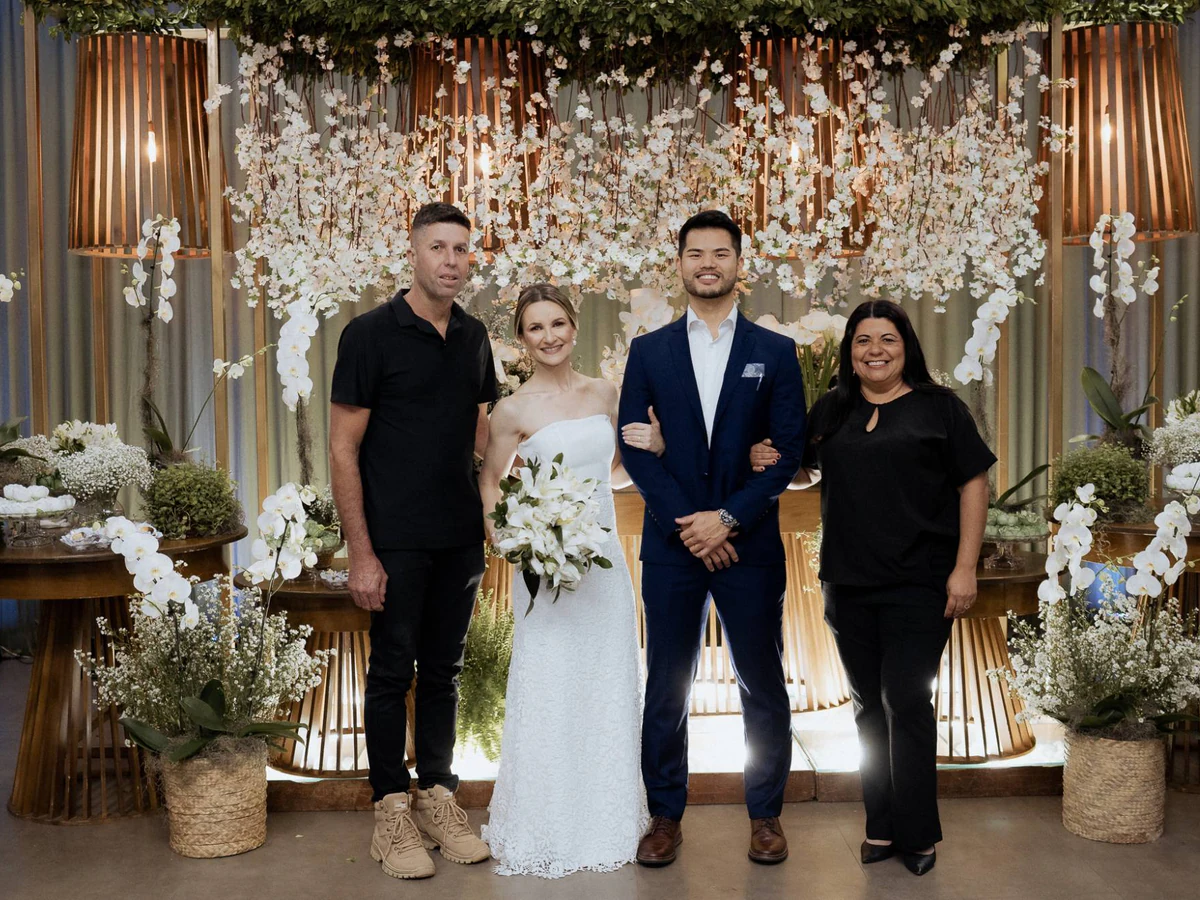 Alline e Anderson ao lado dos noivos diante da mesa principal com cascata de flores brancas e luminárias de madeira ripada