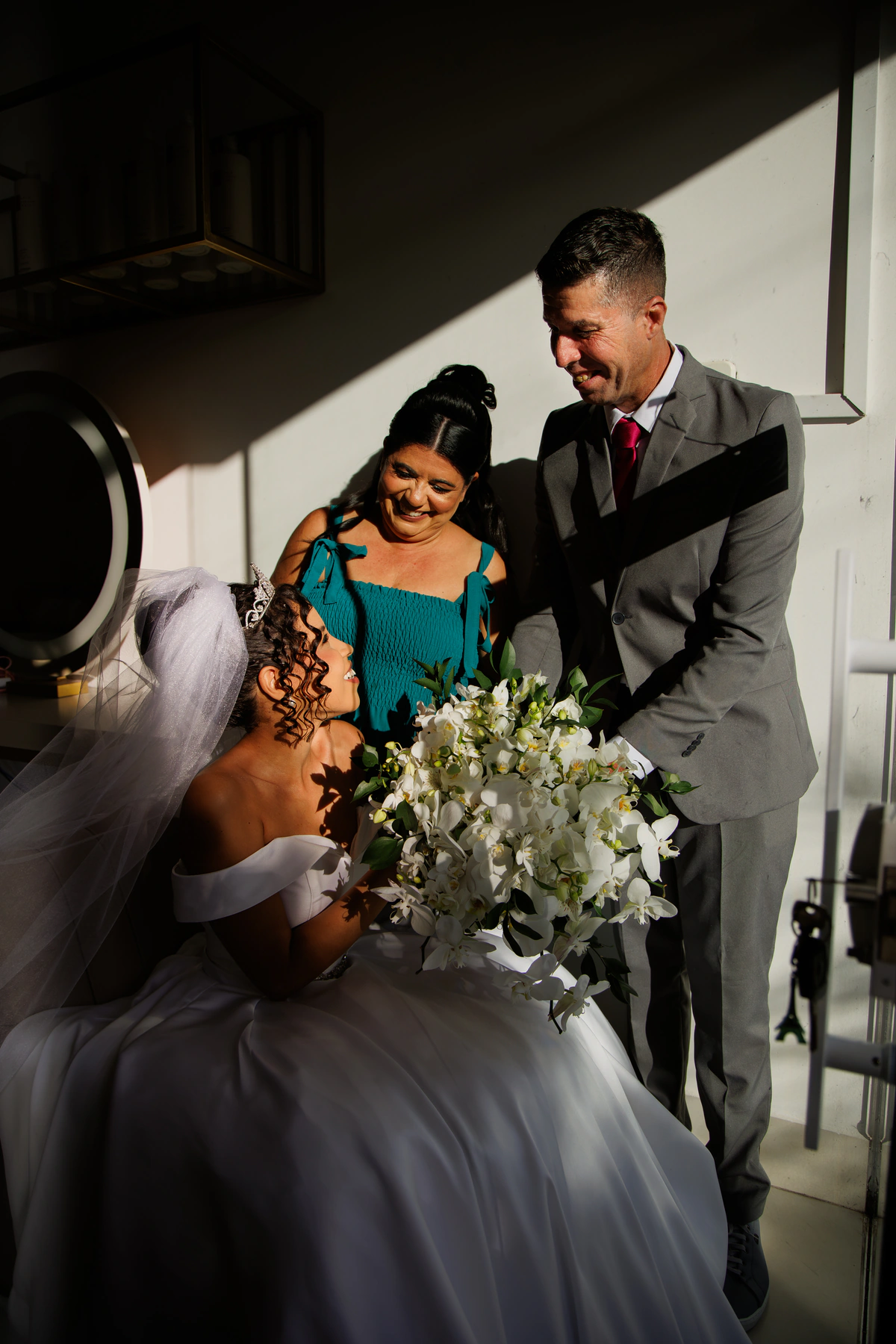 Noiva sentada, de vestido branco, véu e tiara, com o buquê em cascata de orquídeas brancas que o Anderson segura ao lado dela, e a Alline sorrindo ao fundo